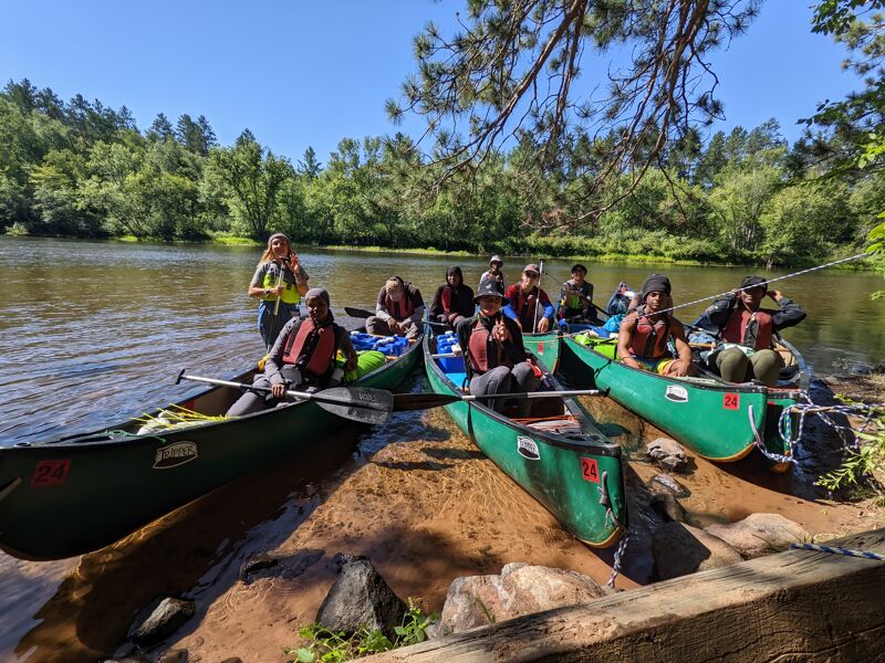 A group of people are gathered on the shore of a lake or river, with several canoes pulled up onto the bank. The people appear to be preparing for or just finishing a canoeing trip. They are wearing casual clothing and life vests. The background features a lush green forest and a clear blue sky, suggesting a sunny day in a natural setting.